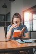 © Satori Studio - Business woman sitting at office desk and writing notes in a notebook while talking on a video call meeting with clients or coworkers on her laptop computer.