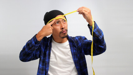 a curious Asian man, dressed in a beanie hat and casual shirt, uses a measuring tape to measure the diameter of his head while standing against white background.