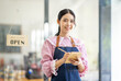 © David - Portrait of happy waitress standing at restaurant entrance. SME entrepreneur young business asian woman attend new customers near door. Smiling small business owner showing open sign in her shop.