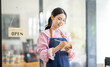 © David - Portrait of happy waitress standing at restaurant entrance. SME entrepreneur young business asian woman attend new customers near door. Smiling small business owner showing open sign in her shop.