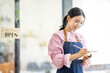© David - Portrait of happy waitress standing at restaurant entrance. SME entrepreneur young business asian woman attend new customers near door. Smiling small business owner showing open sign in her shop.