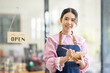 © David - Portrait of happy waitress standing at restaurant entrance. SME entrepreneur young business asian woman attend new customers near door. Smiling small business owner showing open sign in her shop.