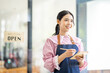 © David - Portrait of happy waitress standing at restaurant entrance. SME entrepreneur young business asian woman attend new customers near door. Smiling small business owner showing open sign in her shop.