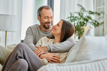 joyful child-free couple embracing and looking at each other on couch in living room, love and joy