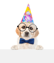 Poster - Golden retriever puppy wearing party cap, tie bow and eyeglasses  looks above empty white banner. isolated on white background