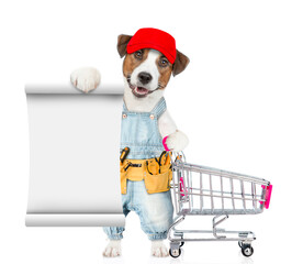 Poster - Funny Jack russell terrier puppy wearing red cap and denim overalls with tool belt shows empty list and holds the shopping cart. isolated on white background