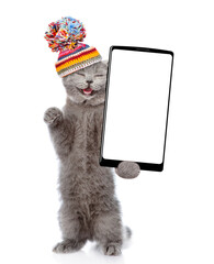 Poster - Happy cat wearing warm knitted hat with pompon holding smartphone with white blank screen in it paw. Isolated on white background