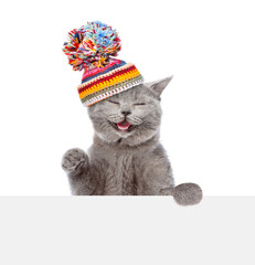 Poster - Happy cat wearing knitted warm woolen hat with pompon looks above empty white board. isolated on white background