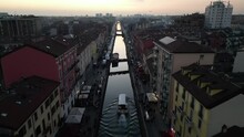 From Above Drone In Motion Of The Navigli District In Milan At Sunset, With A Canal Boat Cruising Down The Waterway And People Walking On The Vibrant Canal-side Walkways