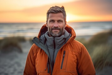 Wall Mural - Portrait of a glad man in his 40s wearing a warm parka against a beautiful beach sunset. AI Generation