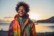 © Markus Schröder - Portrait of a grinning afro-american man in his 20s wearing a vibrant raincoat against a beautiful beach sunset. AI Generation
