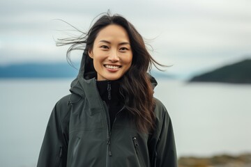 Wall Mural - Portrait of a smiling asian woman in her 20s wearing a windproof softshell against a calm bay background. AI Generation