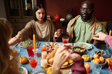 Biracial group of people saying grace holding hands and sitting at thanksgiving table together