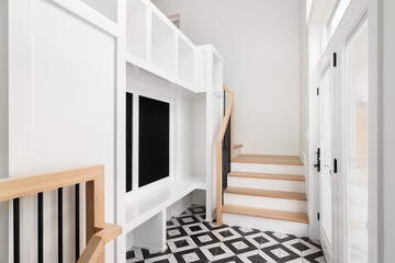 A large foyer with a white storage unit, white bench, white oak stairs and railing, and a black and white pattern tile flooring.