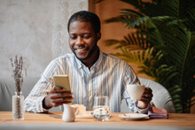 Young African American entrepreneur looking at smartphone screen with smile during communication in video chat while having coffee