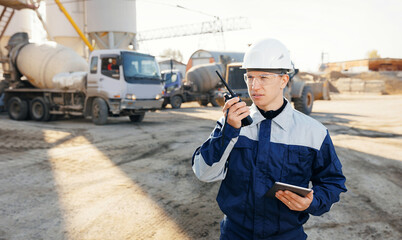 Engineer in uniform and hard hat uses radio walkie to control and coordination for effective work excavator and trucks at cement plant site. Concept Modern optimization of industrial production