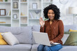 © Tetiana - Smiling African American young woman sitting on sofa at home, talking on video call via laptop, waving and greeting at camera