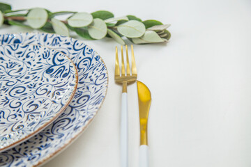 Wall Mural - On the side, two serving plates with blue patterns on a white background stand one on one, part of the plates can be seen, gold-white appliances are located on the side, a green branch of plants lies