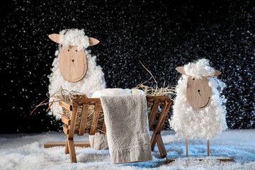 Wall Mural - Toy sheep and wooden manger with dummy of baby on snow against black background. Concept of Christmas story