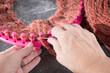 Soft, wool, yarn in a burnt orange color.  Hands using a hook tool on a pink, round knitting loom with both knit and pearl stitches.