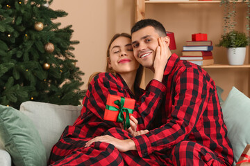 Canvas Print - Young couple wearing plaid pajamas with gift in living room decorated for Christmas