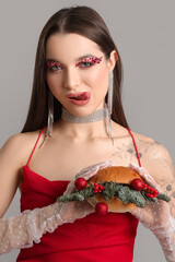 Wall Mural - Beautiful young woman holding burger with Christmas balls and fir branches on grey background