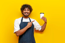 Young Indian Guy Barista In Apron Pointing At Paper Cup Of Coffee On Yellow Isolated Background