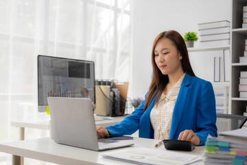 Asian female employee using calculator and laptop to do financial math on tax calculation table Accounting report, company financial statistics and analytical research ideas at the office