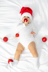 Wall Mural - Full length top view portrait of a newborn baby. Cute Caucasian baby 6-7 months old in red Santa hat and white suit costume lying on a cozy bed with Christmas ball in hand.