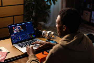 Wall Mural - Delighted serious handsome attractive African American professional videographer, using his phone to capture behind-the-scenes footage while editing videos on laptop.