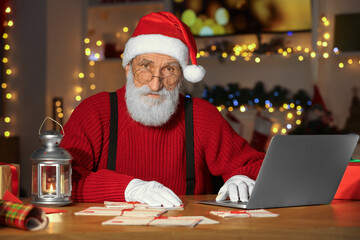 Canvas Print - Santa Claus at his workplace. Letters and laptop on table in room decorated for Christmas