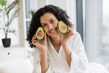 Beautiful calm multiracial female in robe holding two avocado halves in hands near cheek in bedroom interior. Young African American curly woman appreciating green avocado for great skin benefits.