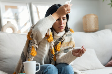 Tired woman with a cold taking her fever while lying with a blanket on the couch at home