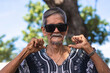 A cool hippie granddad pointing to the camera pointing to himself with both thumbs with confidence. Wearing shades and boho style shirt. Hanging out at the park.
