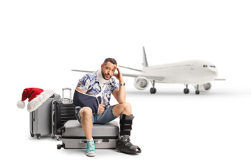 Canvas Print - Man with an injured arm and leg sitting on suitcases in front of an airplane, travelling for christmas