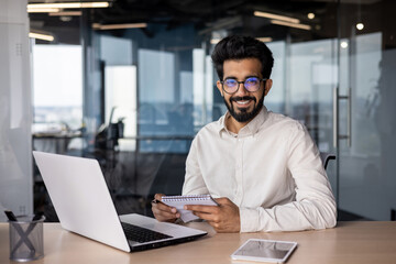 Wall Mural - Portrait of a young Indian man sitting in the office at the table, working on a laptop, holding a notebook, smiling and looking at the camera