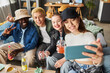 High angle shot of beaming multi-ethnic young people taking selfie during party in living room, focus on brunette girl in cap holding smartphone