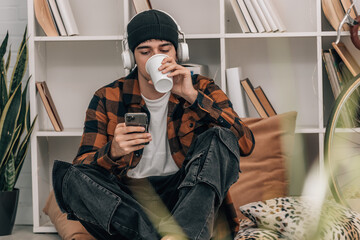 Poster - young man at home drinking while looking at phone with headphones