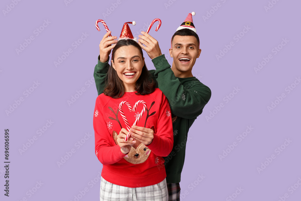 Happy young couple in Christmas pajamas and with candy canes on lilac background