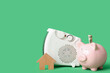 Electric heater with piggy bank, money and wooden house on green background. Concept of heating season