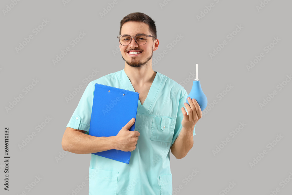 Young doctor in eyeglasses with clipboard and enema on grey background