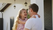 Smiling American Family Play Outdoors Near The House On A Warm Summer Day. A Joyful Father Holds Daughter In Arms And Spins Her Around. Mom, Dad And Daughter Are Hugging. Husband Kisses A Wife. The