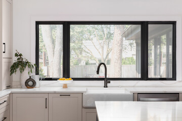 Wall Mural - A kitchen detail with a black faucet on a white farmhouse sink in front of black framed windows, brown cabinets, and white marble countertops.