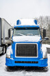 Front of the blue big rig semi truck covered with snow and ice standing on the truck stop winter parking lot beside another semi truck with semi trailer