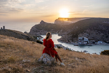 Wall Mural - Woman sunset sea mountains. Happy woman siting with her back on the sunset in nature summer posing with mountains on sunset, silhouette. Woman in the mountains red dress, eco friendly, summer