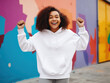 African-American teenage girl wearing a blank white hoodie with no print or logo standing in front of colorful wall, raising hands in excitement, apparel mockup photo, street fashion
