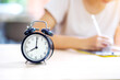 Selective focus of alarm clock on table near young female student in casual cloth doing homework at her school. concept of time management.