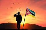 Fototapeta  - Silhouette of a soldier with the Mozambique flag stands against the background of a sunset or sunrise. Concept of national holidays. Commemoration Day.