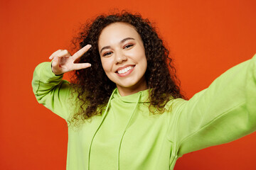 Wall Mural - Close up young woman of African American ethnicity wear green hoody casual clothes do selfie shot pov mobile cell phone cover eye with v-sign isolated on plain red orange background Lifestyle concept