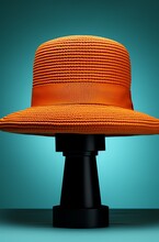 A Bright Orange, Woven Hat Placed On Top Of A Black Stand. The Background Is Gradient Teal Blue, Transitioning From Dark At The Corners To Light In The Center, Creating A Spotlight Effect.